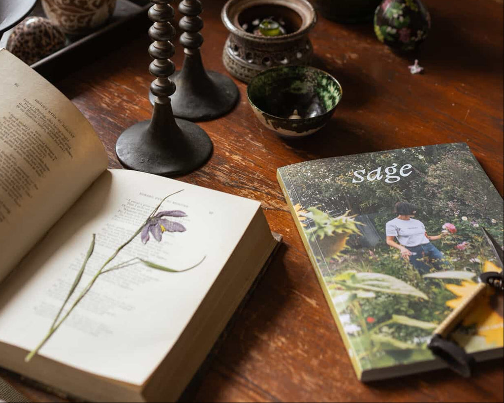 A styled tabletop scene with a dark wooden table holding a book with an open page and pressed flower, a magazine or booklet titled sage, two black candlesticks with green candles, small ceramic vases, bowls, and a green vase with pale yellow flowers.